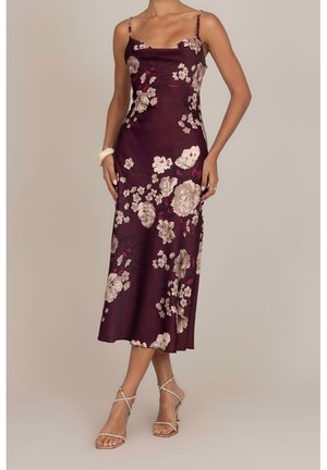 Vrouw draagt een mouwloze, donkerpaarse midi-jurk met bloemenprint, dunne bandjes en beige sandalen met hoge hakken en bandjes, staand tegen een effen achtergrond.