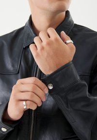 Giacca in pelle nera con zip frontale, polsini con bottoni e una texture morbida. Mani che mostrano un anello d'argento, evidenziando la lavorazione dettagliata.