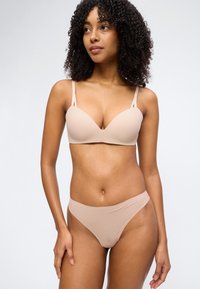 Γυναίκα με σγουρά μαλλιά που φοράει σουτιέν σε γυμνό χρώμα και εσώρουχα με ψηλό κόψιμο στο ίδιο χρώμα, στέκεται μπροστά σε απλό φωτεινό φόντο.