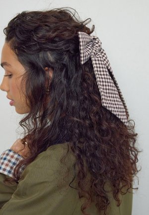 Curly brown hair styled with a checkered fabric bow in shades of brown and white. The background is neutral, highlighting the hair accessory.