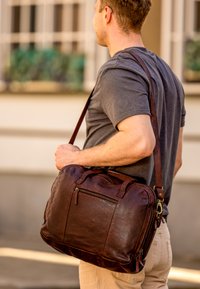Man wearing gray t-shirt and beige pants carrying a brown leather shoulder bag outdoors in front of a building.
