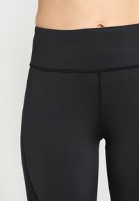 Detailní záběr pasu osoby, která má na sobě černé, hladké legíny s vysokým pasem, na jednobarevném světlém pozadí.
