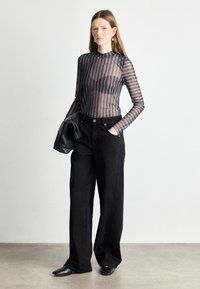 Jeans neri a gamba larga abbinati a un top trasparente a righe in blu scuro e marrone. Il modello tiene una borsa nera. Tessuto morbido, design aderente.