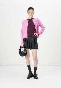Vrouw in een bril, met een roze cardigan, bordeaux top, zwarte geplooide rok, die een zwarte handtas vasthoudt en met haar hand op de heup tegen een witte achtergrond staat.