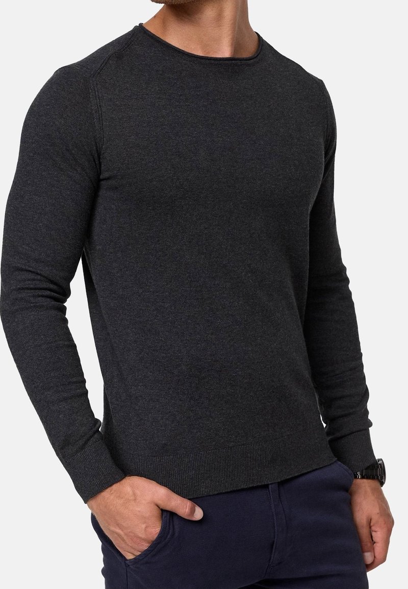 Homme portant un pull cintré à manches longues gris foncé et un pantalon bleu marine, une main dans la poche et une montre-bracelet au poignet gauche.