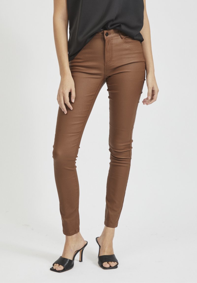 Pantalon marron ajusté en matériau lisse, taille haute, jambe slim et longueur raccourcie, associée à des sandales à talons noires.