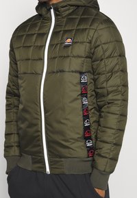 Man in een olijfgroene gewatteerde jas met witte rits, capuchon en ellassee logo patches op de borst en zijstrook.