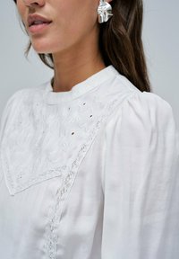 Žena nosi bijelu bluzu s čipkastim detaljima i cvjetnim vezom, uparenom s velikom, sjajnom srebrnom naušnicom.