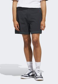 Schwarze Shorts mit türkisfarbenen Nähten, aus weichem Material. Mit einem elastischen Bund und seitlichen Taschen, getragen mit weißen Socken und Turnschuhen.