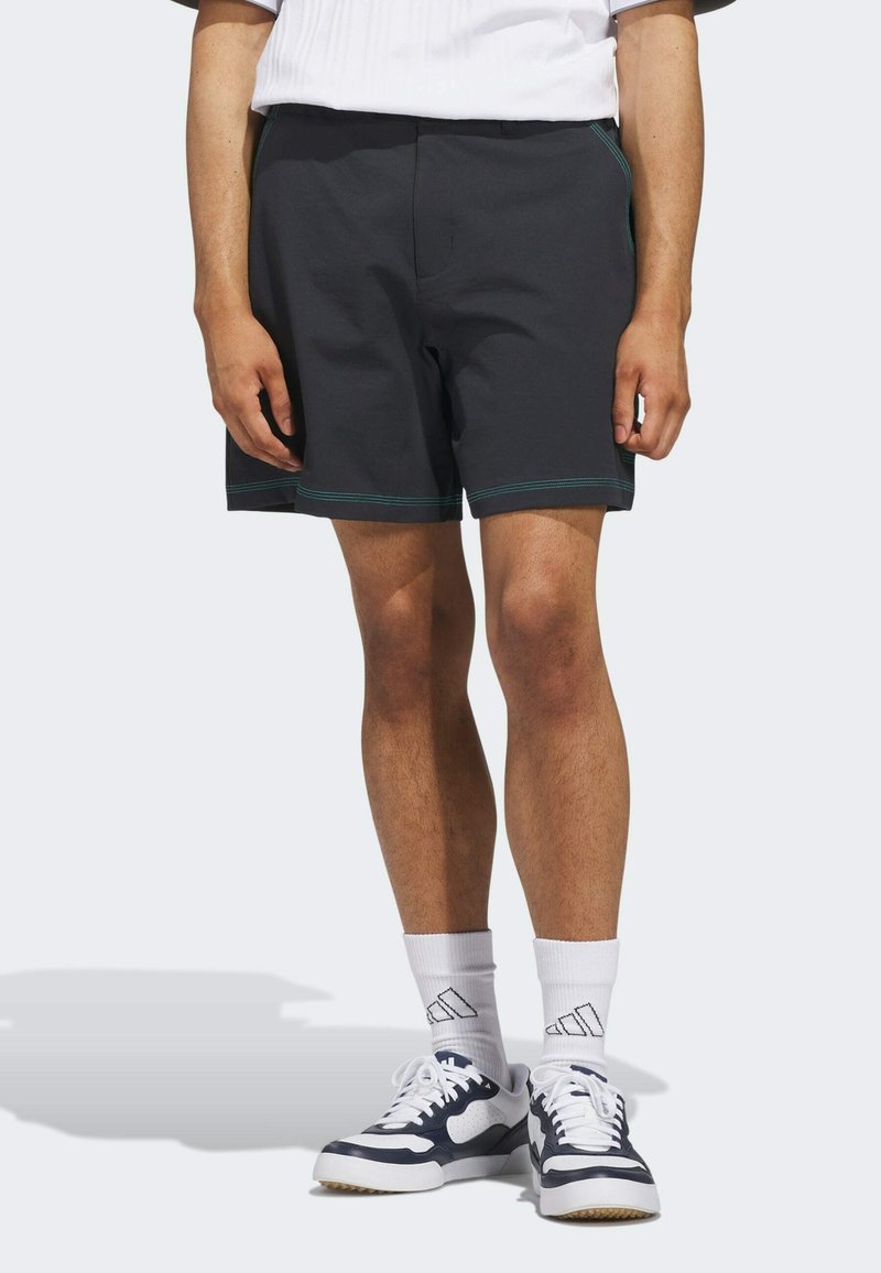 Schwarze Shorts mit türkisfarbenen Nähten, aus weichem Material. Mit einem elastischen Bund und seitlichen Taschen, getragen mit weißen Socken und Turnschuhen.
