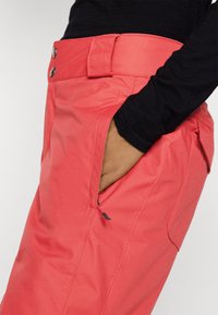 Pantalons couleur corail en tissu durable avec une poche zippée, deux boutons à l'avant et une taille ajustée avec passants de ceinture.