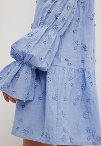 Svetlo modra vezenina obleka s napihnjenimi rokavi, ki vključuje cvetlične vzorce in teksturirane podrobnosti. Mehka tkanina z nabranimi akcenti.