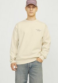 Sudadera beige de manga larga y cuello redondo, con un pequeño detalle de texto en el pecho. Combinada con jeans azules.