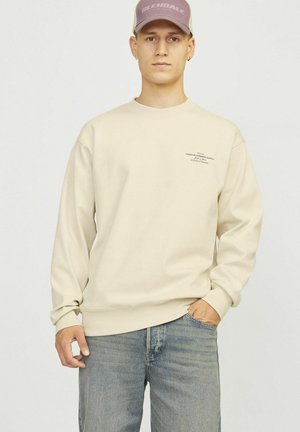 Sudadera beige de manga larga y cuello redondo, con un pequeño detalle de texto en el pecho. Combinada con jeans azules.