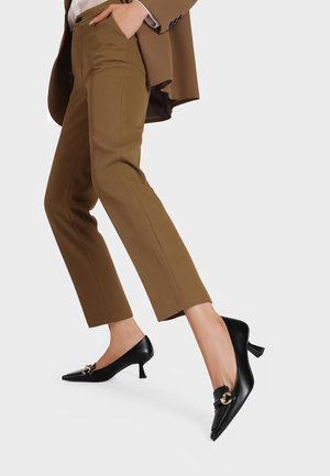 Scarpe nere in pelle con tacco alto, dalla punta affilata, dotate di dettagli in oro e un piccolo tacco quadrato, abbinate a pantaloni marroni su misura.