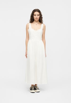 Jeune femme aux cheveux bouclés portant une robe midi blanche sans manches et des baskets noires, debout devant un fond gris uni.