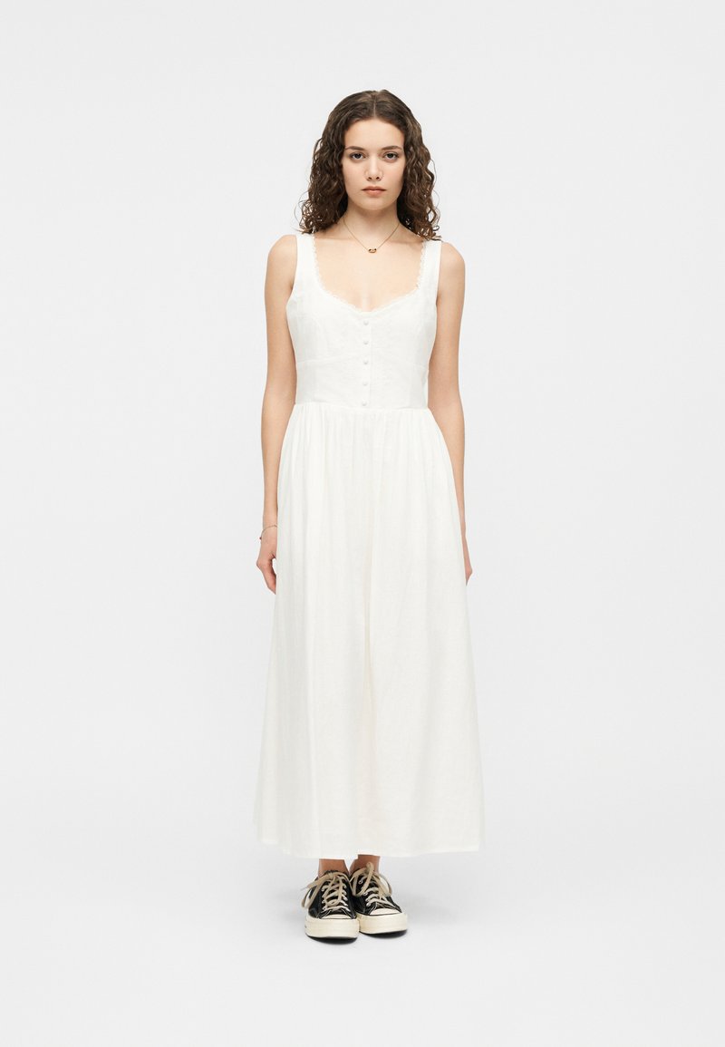 Νέα γυναίκα με σγουρά μαλλιά, ντυμένη με ένα λευκό αμάνικο μεσαίο φόρεμα και μαύρα αθλητικά παπούτσια, στέκεται μπροστά σε ένα απλό γκρι φόντο.