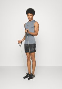 Graues ärmelloses Sporttop, schwarze gemusterte Shorts und schwarze Sneakers. Hält Widerstandsbänder und zeigt einen muskulösen Körperbau.
