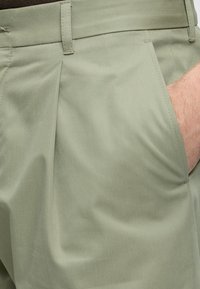 Pantalon vert clair avec une main insérée dans la poche avant droite, montrant des passants de ceinture et des détails de tissu plissé.