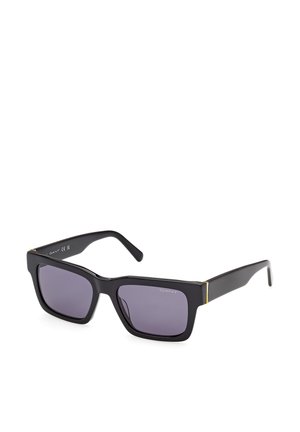 Lunettes de soleil noires rectangulaires avec des montures épaisses, des verres foncés et de subtils accents dorés près des charnières, inclinées sur un fond blanc.
