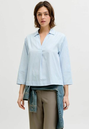 Žena nosící světle modrou blůzu s pruhy a 3/4 rukávy, k níž má uvázanou džínovou bundu přes béžové kalhoty.