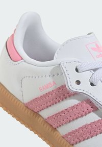 Biały tenisówka z różowymi paskami zamszowymi, wyposażona w białe sznurowadła i jasnoróżowy obcas. Na boku znajduje się znak "SAMBA" w kolorze różowym. Gumowa podeszwa.