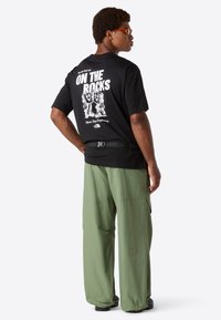 Muž v černém tričku s potiskem "On The Rocks", se zelenými širokými kalhotami, oranžovými slunečními brýlemi a černým páskem, stojící bokem.