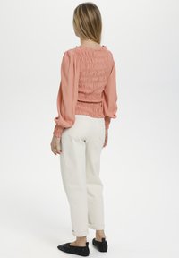 Persikofärgad blus med smock och långa puffärmar, som har en subtil textur, i kombination med vita byxor med uppvikt ärm och svarta slip-on skor.