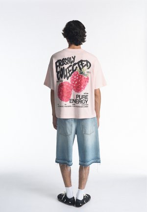 Personne portant un T-shirt rose avec de grands motifs de framboise et du texte, associée à un short en jean clair et des baskets noires, debout de dos.