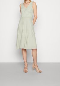 Ljust grön klänning med spets-bodice och A-linjeformad kjol, med en V-ringning och veckad textur. Kombinerad med ljusa strappy högklackade skor.