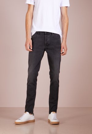 Homme portant un jean slim noir, un t-shirt blanc et des baskets blanches, debout devant un fond beige uni.