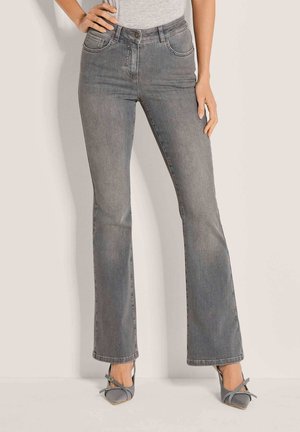 Femme portant un jean flare gris taille haute et des talons pointus gris avec des brides à la cheville, debout, une main sur la hanche.