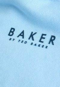 Svijetloplava tkanina s crnim tekstom: "BAKER" u podebljanom fontu, praćena "by TED BAKER" u manjoj tipografiji. Glatka tekstura, ležeran stil.