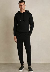 Hoodie negru cu șnururi, pantaloni de trening asortați și adidași negri. Material neted, design ajustat, manșete și talie ribbate.
