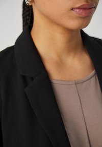 Blazer negro con solapas con muesca, de tela texturizada, usado sobre una camiseta beige de escote redondo, que destaca un diseño limpio y minimalista.