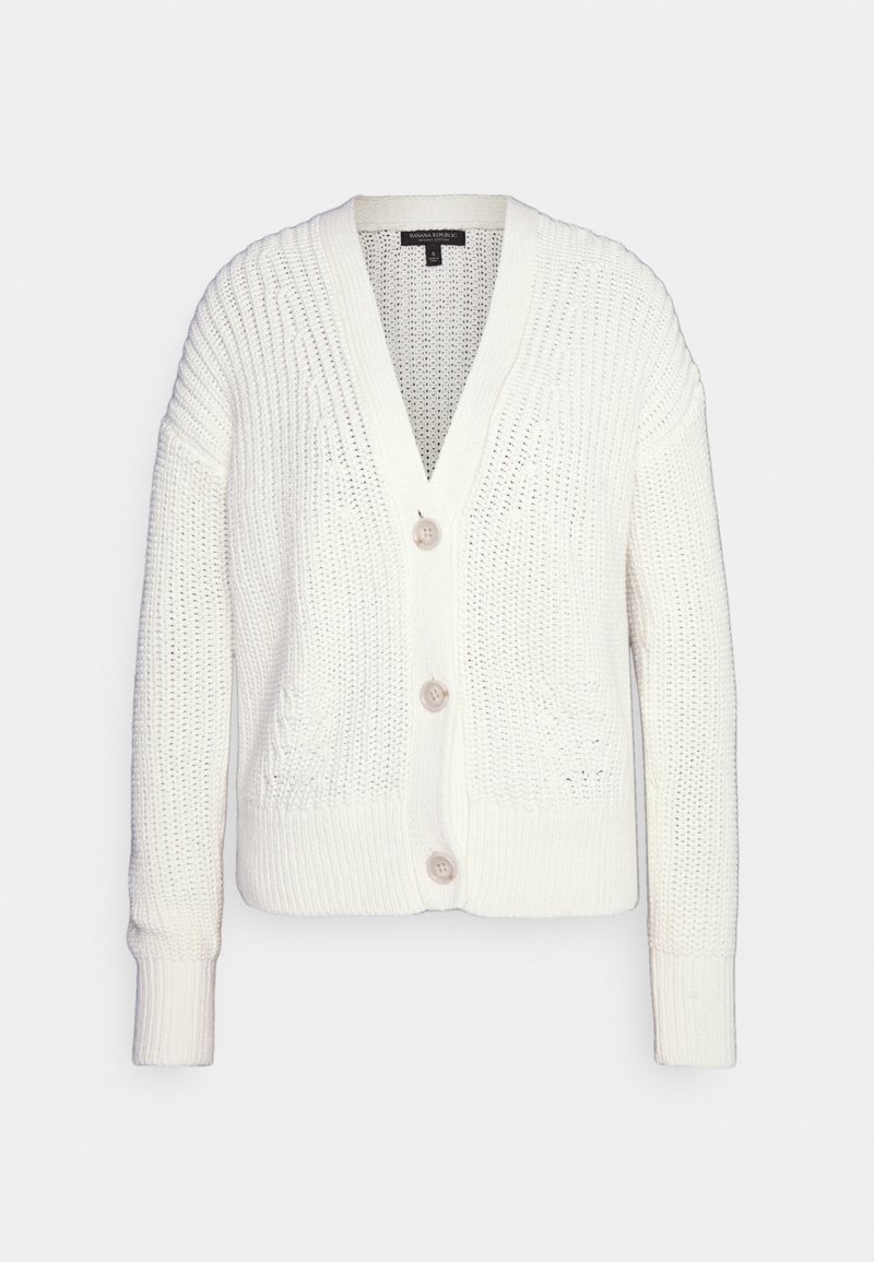 Valkoinen neule cardigan, jossa on V-pääntie, kolme suurta nappia, pitkät hihat ja ribbireunukset. Ominaisuuksiin kuuluvat avoin neulos ja kaksi etutaskua.