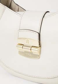 Kremowa skórzana torebka o zaokrąglonym kształcie, z złotym zapięciem i logo marki. Gładka faktura z widocznymi szwami i akcentami.