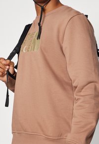 Sudadera marrón hecha de tejido suave, con un logo verde en el pecho, cuello redondo y puños acanalados para un ajuste ceñido.