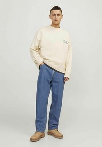 Béžová mikina s kulatým výstřihem a potiskem, doplněná modrými volně střiženými džínami a hnědými šněrovacími botami. Jednobarevný a ležérní design.