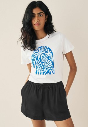 Weißes T-Shirt mit blauem floralem Grafikdesign, das Blattmuster zeigt, kombiniert mit schwarzen Shorts mit elastischem Bund. Glattes Baumwollmaterial.