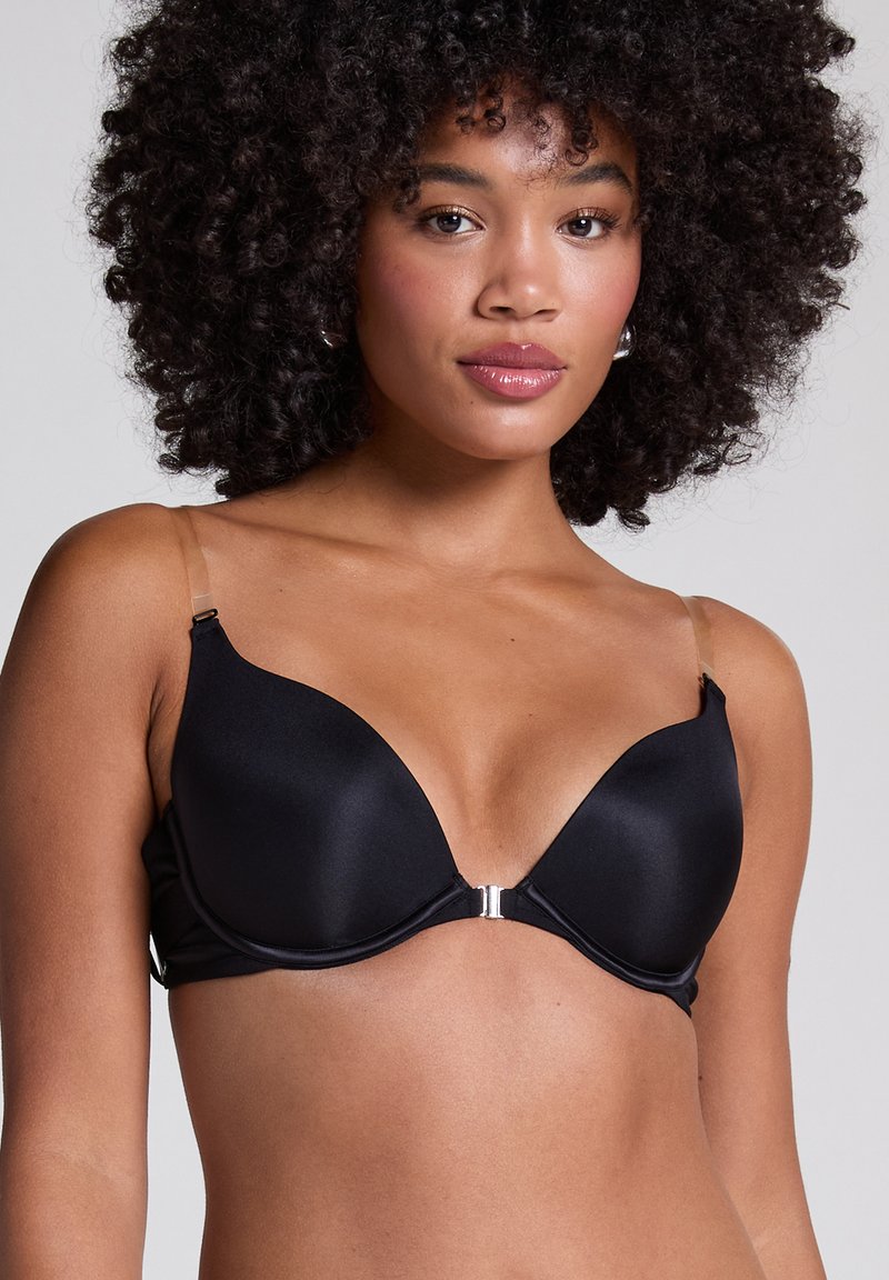 Frau mit lockigem Haar, die einen schwarzen BH mit Frontverschluss und transparenten verstellbaren Trägern vor einem schlichten grauen Hintergrund trägt.