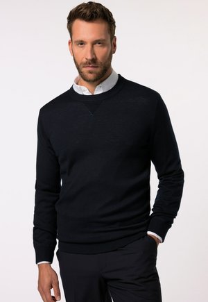 Pull en tricot navy avec un col rond, des poignets et un ourlet côtelés, et un accent texturé en forme de V au niveau de la poitrine. Porté par-dessus une chemise à col blanc.