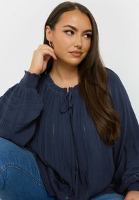 Blusa blu scuro in tessuto testurizzato, con scollo a chiave legato, maniche a sbuffo e dettagli in pizzo lungo le cuciture.