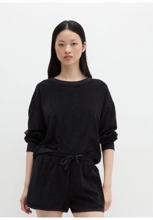 Młoda kobieta z długimi czarnymi włosami, ubrana w czarny długi rękaw i dopasowane czarne szorty na jednolitym białym tle.