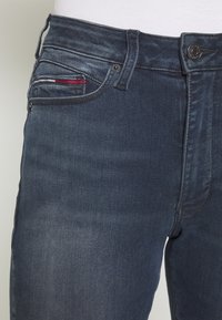 Pantalones vaqueros de mezclilla azul oscuro con un diseño clásico, que cuentan con un bolsillo frontal con costuras de acento en rojo, blanco y azul, y cierre de botón metálico.