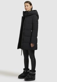 Sort puffervinterjakke med høj krave, lynlås og trykknaplukning. Har en firkantet silhuet og bæres med sorte leggings og støvler.