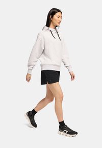 Hellgrauer oversized Hoodie mit schwarzen Kordeln, kombiniert mit schwarzen Sportshorts. Schwarze Sneaker mit weißer Sohle und Knöchelsocken.