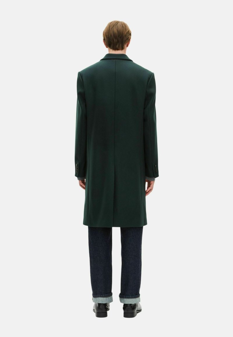 Long dark green coat with a structured shape, wide lapels, and visible seams, paired with dark jeans and black shoes.