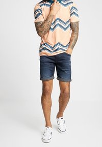 Kortärmad skjorta i persiko och blått zigzag-mönster, matchad med mörka denimshorts som är uppvikta vid fållen, kompletterad med vita sneakers.