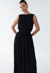 Schwarzes Maxikleid mit texturiertem, plissiertem Stoff. Ärmelloses Design, tailliert und mit einem Kordelzug mit goldenen Akzenten. Vor hellem Hintergrund dargestellt.